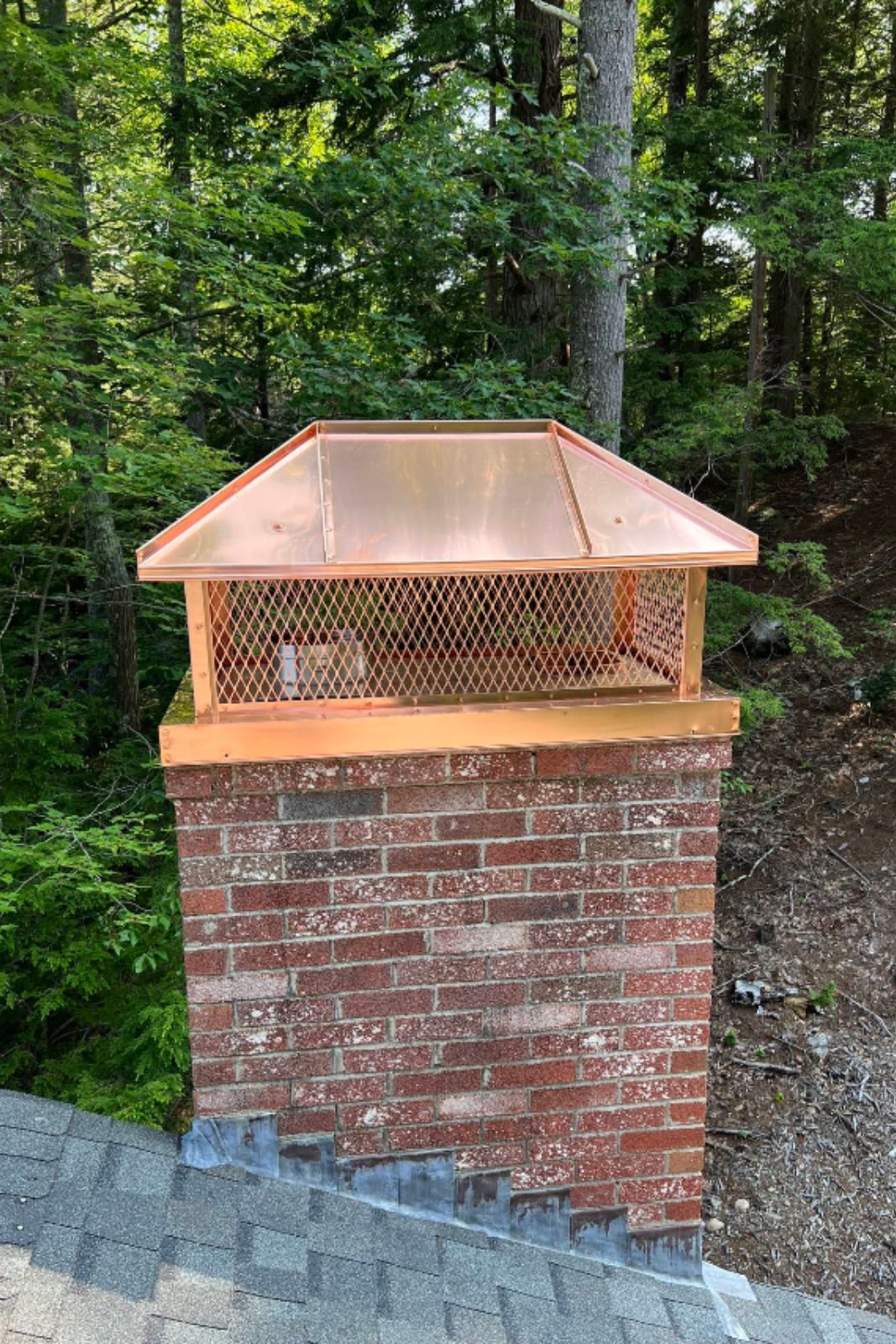 Copper chimney cap with mesh on brick chimney, forested backdrop; location Sanford, ME.