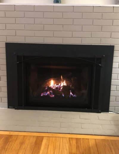 Gas fireplace insert installation in Cape Elizabeth, ME featuring black arched surround and traditional brick fireplace by Frechette Chimney Sweeping.